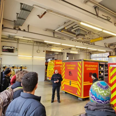 Exkursion zu einer Feuerwache - vorne Teilnehmer der Veranstaltung, in der Mitte des Bildes steht ein Feuerwehrmann und im Hintergrund sind mehrere Fahrzeuge der Dresdener Feuerwehr zu sehen